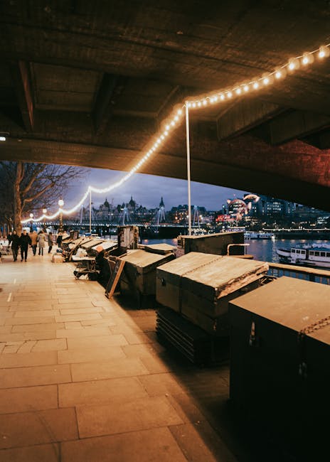 A view of a bridge spanning a river with classical-style lamp posts along its balustrade, leading toward a modern, glass-covered skyscraper that leans sharply to the left in the background, with the s